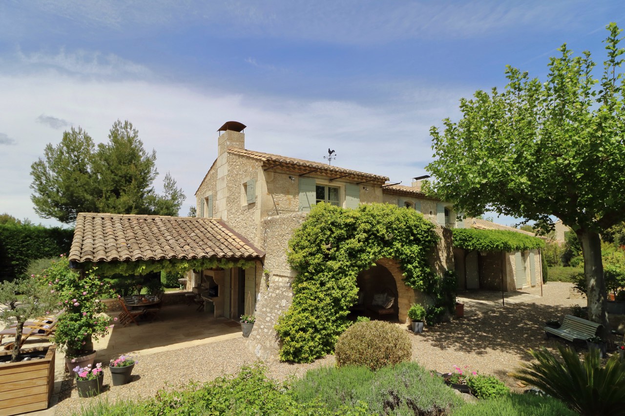 A traditional stone house with a tiled roof, surrounded by lush greenery and a clear blue sky. the yard features a graveled area with outdoor seating and potted plants.